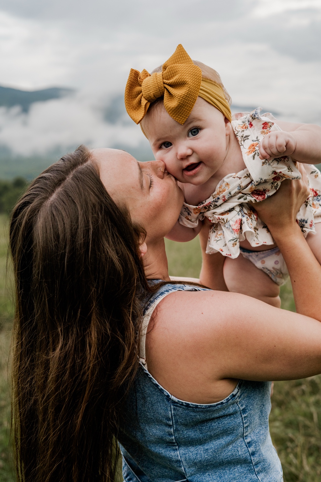 A mother kissing her smiling baby, wearing a floral outfit and mustard bow, with misty mountains behind them